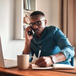 Man by his computer and notepad taking notes while on a phone call