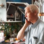 Older man looking out the window of his home's kitchen while on a phone call