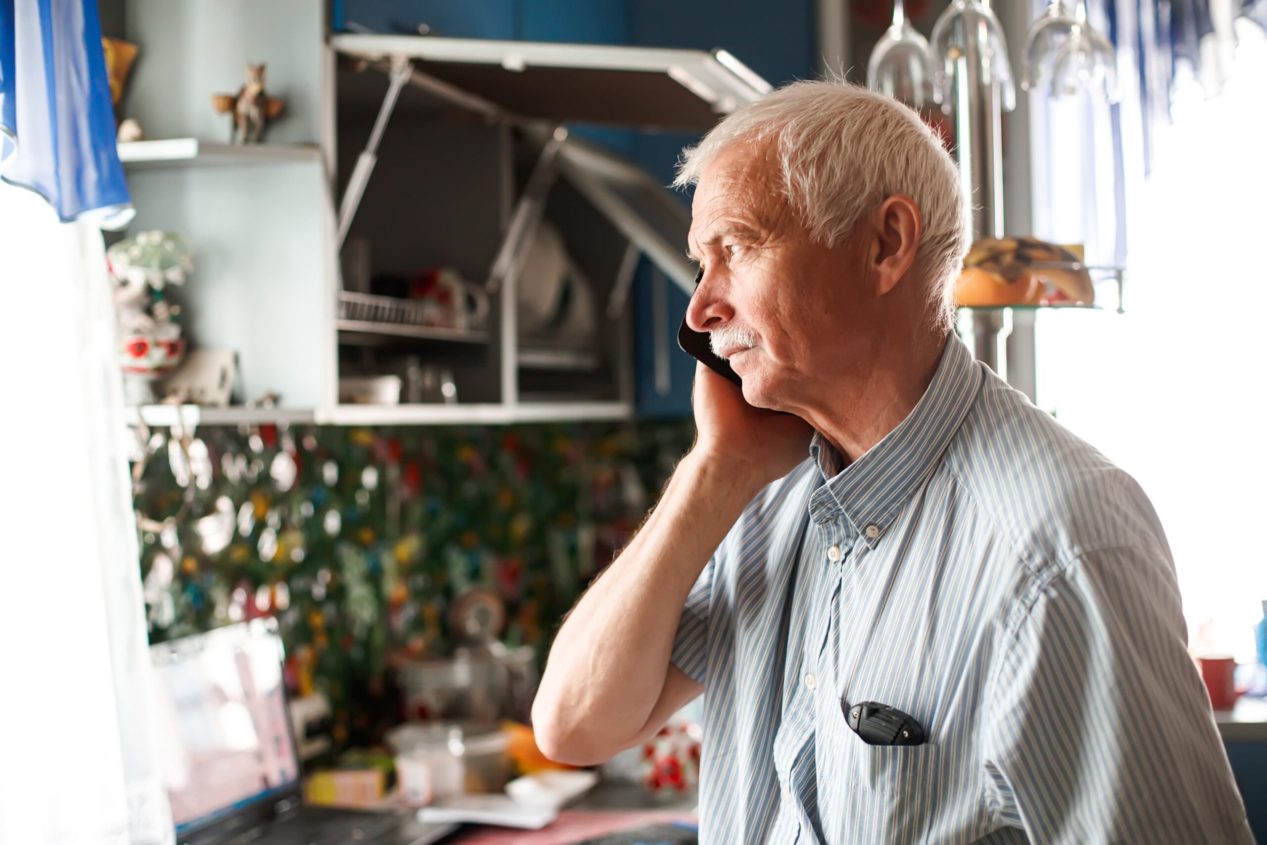 Older man looking out the window of his home's kitchen while on a phone call