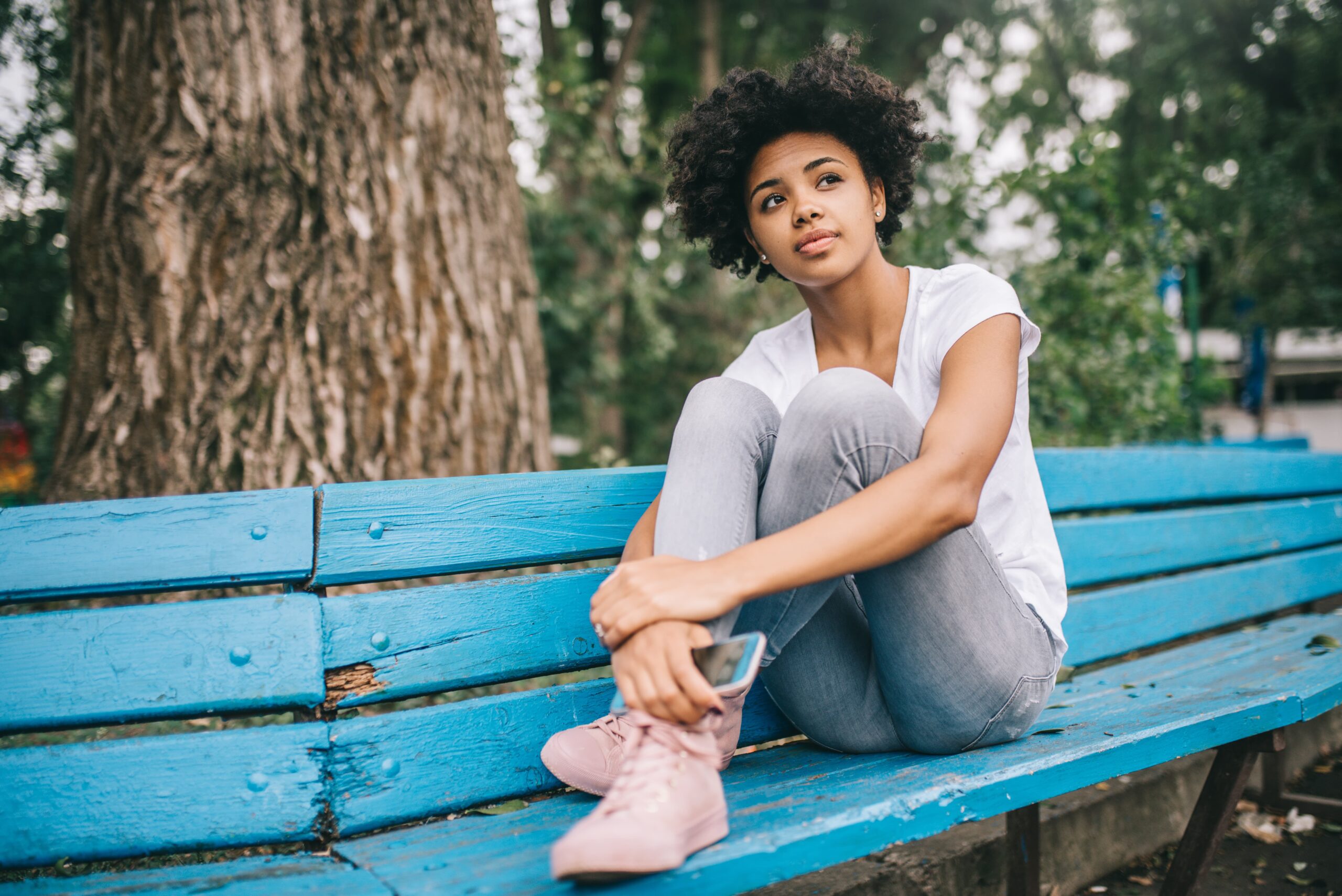 Teenage girl sitting on bench in a city with phone in her hand and looking beyond the camera