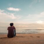 Man sitting on beach looking at the water facing away from camera