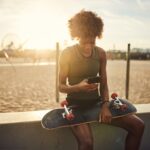 Young woman texting on her phone while sitting at the beach with her skateboard