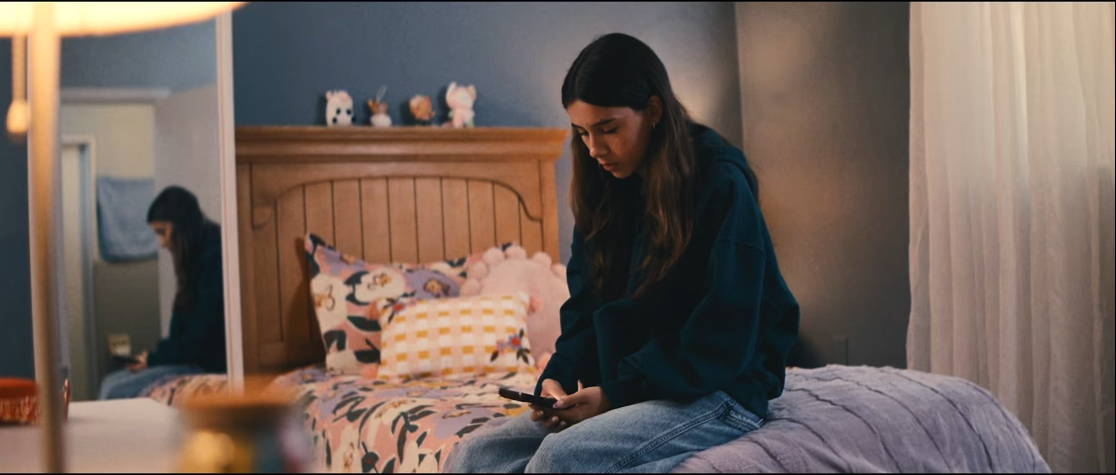 young girl with long brown hair sitting on the side of a bed looking at a cell phone